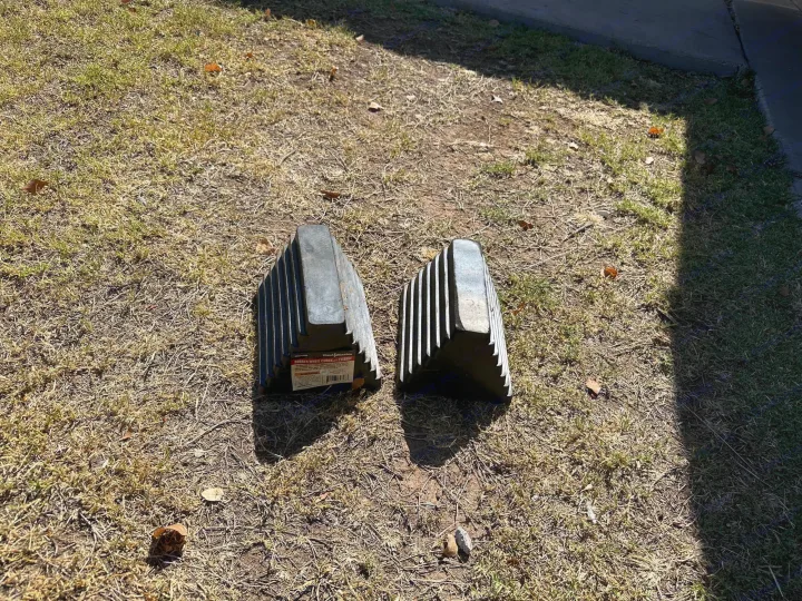 Two dark rubber wheel chocks sit on a dry, grassy lawn, vital for securing an rv rental and preventing unwanted movement.. Coleman Lantern LT 17B 2023