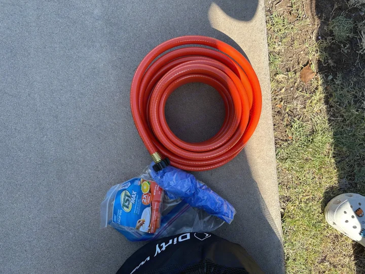 Essential RV rental supplies: Camco TST MAX toilet treatment and a red water hose on concrete next to grass and a shoe.. Coleman Lantern LT 17B 2023