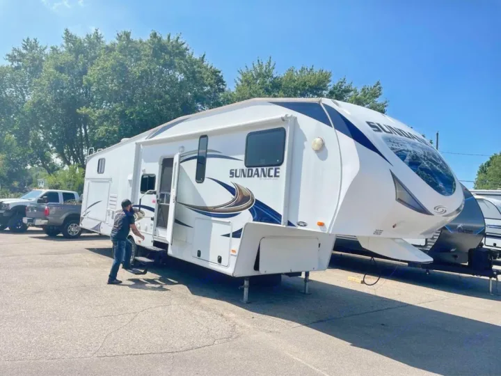 A large white and blue Sundance fifth-wheel RV is parked outdoors, with a person nearby. Explore this RV for rental.. Heartland RVs Sundance 2012