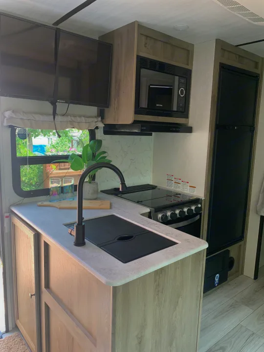 Compact RV rental kitchen featuring microwave, fridge, stove, sink, and dual TVs. Modern light wood & gray tones.. Forest River Surveyor Legend 2022