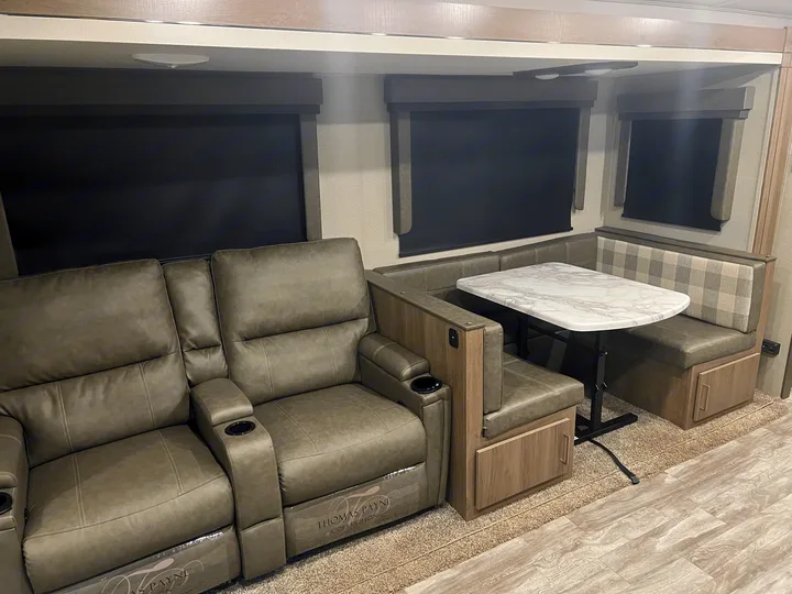 RV rental interior: two leather recliners with cup holders, alongside a dining booth with a white marble table and plaid cushions.. Grand Design Imagine 2022