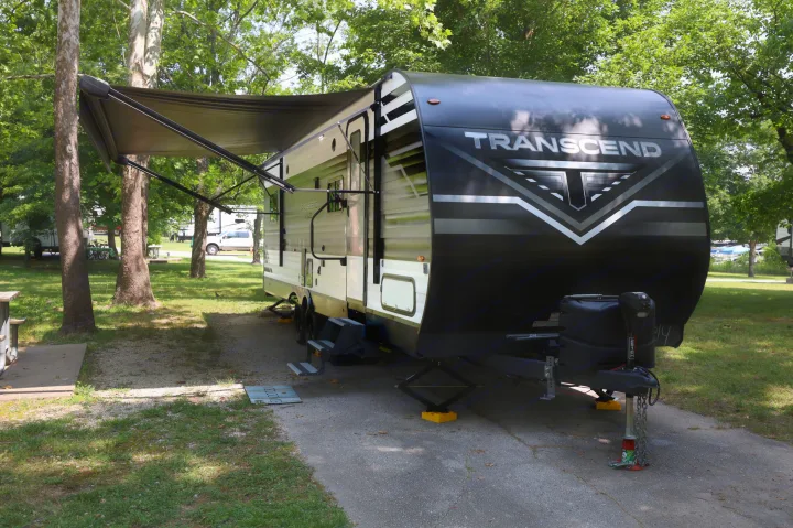 A black and white Transcend travel trailer, often used as an RV rental, is parked with its awning extended at a shaded campsite.. Grand Design Transcend Xplor 2022