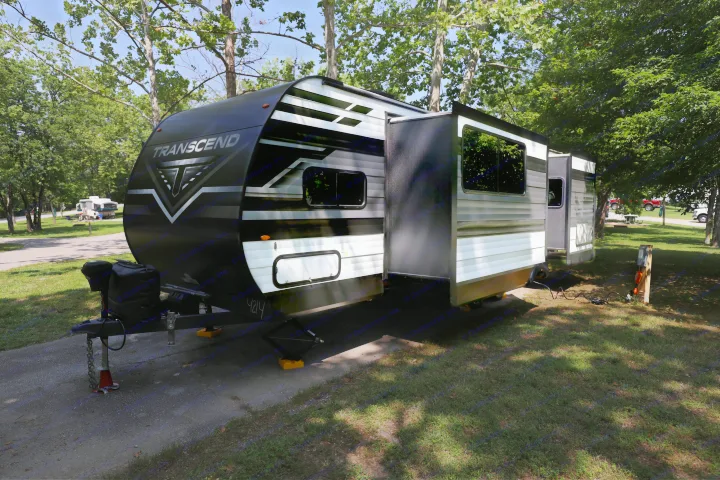 A Transcend travel trailer RV, perfect for a rental, is set up at a shaded campsite, showcasing its extended slide-out.. Grand Design Transcend Xplor 2022