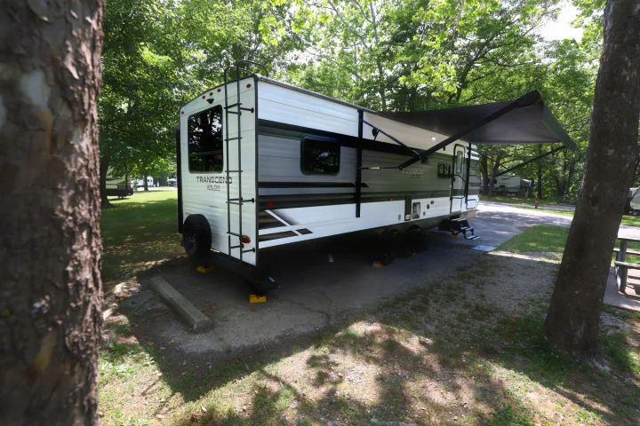 Enjoy a peaceful rv rental experience! A white and gray travel trailer with awning extended at a shaded campground campsite.. Grand Design Transcend Xplor 2022