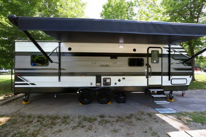 This modern RV rental travel trailer is set up with an extended awning and steps down at a shaded campsite.. Grand Design Transcend Xplor 2022
