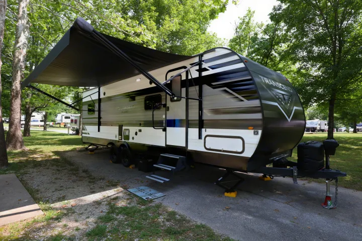 A modern RV rental with an extended awning sits on a gravel campsite pad, surrounded by lush green trees.. Grand Design Transcend Xplor 2022