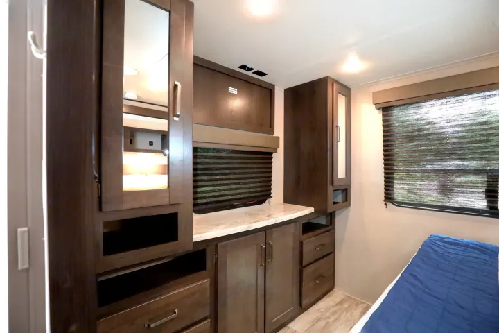 This RV rental bedroom offers dark wood cabinetry, a mirror, window with blinds, and a cozy bed.. Grand Design Transcend Xplor 2022