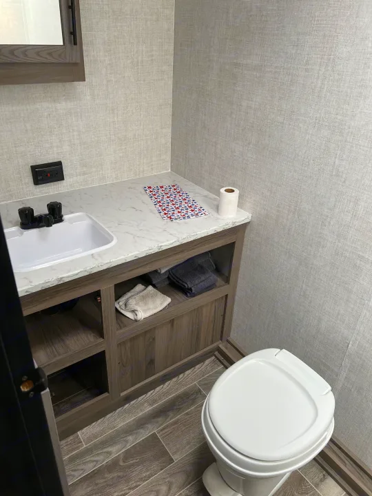 Modern RV rental bathroom featuring a sink, toilet, white counter, and wood-look floor. Shelves hold towels.. Gulf Stream Ameri-Lite 2022