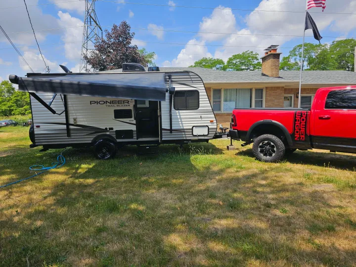 A red Dodge Power Wagon pickup truck is hitched to a white Pioneer travel trailer RV, parked on a sunny lawn for rental.. Heartland RVs Pioneer 2020