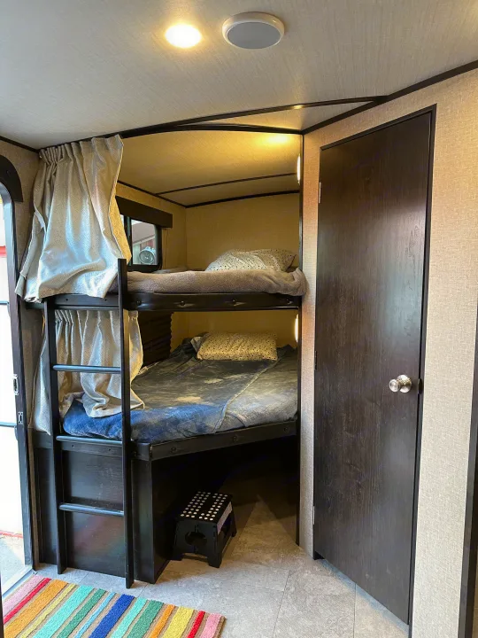 Cozy RV rental interior: stacked bunk beds with bedding, privacy curtains, and a dark door. Ideal compact sleeping area.. Grand Design Imagine 2020