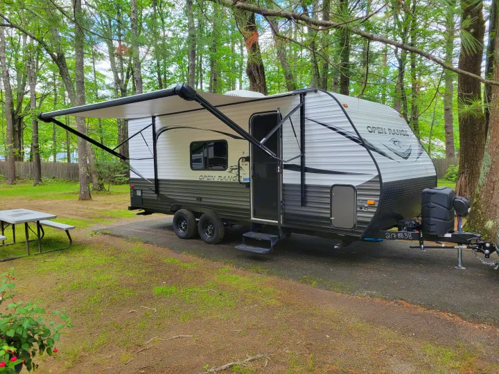 RV rental travel trailer with an extended awning parked at a green, wooded campsite with a picnic table.. Highland Ridge RV Open Range 2021