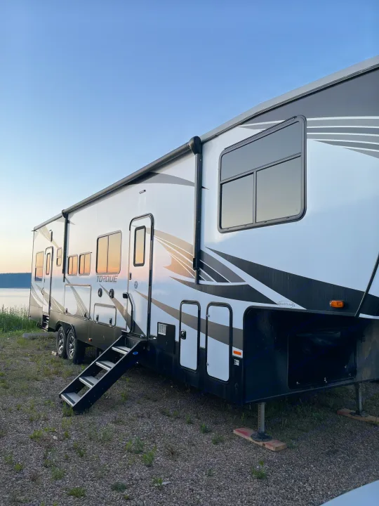 Scenic lakeside RV rental: A white and black Torque fifth wheel camper parked on gravel, its windows reflecting the sunset.. Heartland Torque 2020