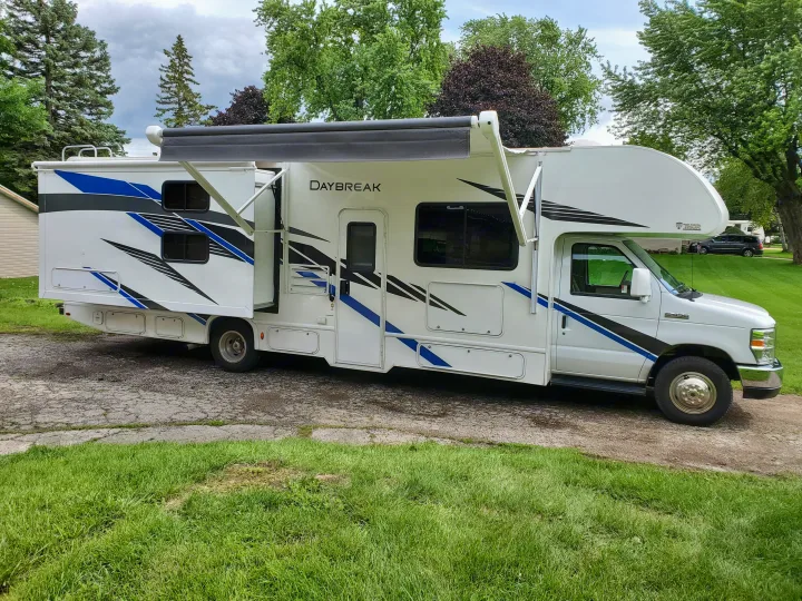 White Class C RV rental with extended awning, blue/black graphics, parked on pavement by green grass.. Thor Motor Coach Daybreak 30D 2020