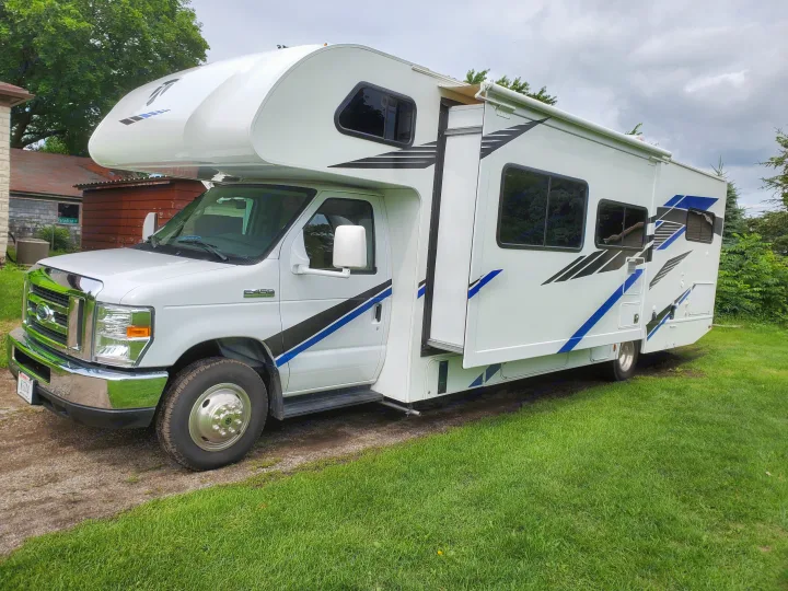 White Ford Class C RV rental with blue/black graphics and a slide-out, parked on gravel next to green grass.. Thor Motor Coach Daybreak 30D 2020
