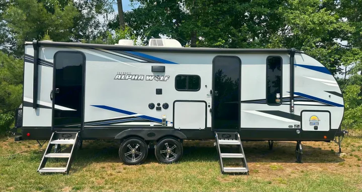 This grey Cherokee Alpha Wolf travel trailer RV rental, with two entrance doors & steps, is parked in nature.. Forest River Cherokee Alpha Wolf 2022