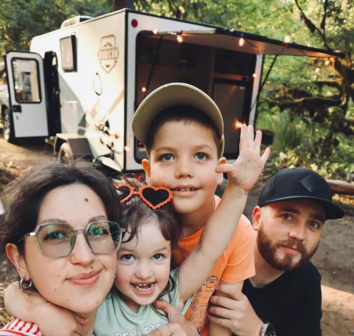 A happy family poses with their Rubicon RV rental, illuminated by string lights, on a memorable camping adventure.. Dutchmen Coleman Rubicon 2023