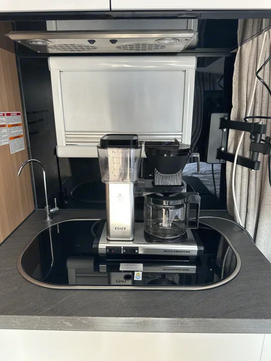 A well-equipped RV rental kitchen featuring a Moccamaster coffee maker, sink, and overhead range hood.. Black Series Black Series Trailer 2022