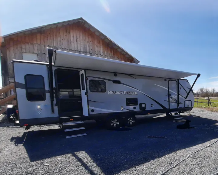 Grey Shadow Cruiser RV rental with awning extended, parked on gravel next to a barn, with a black dog resting.. Cruiser RV Shadow Cruiser 2019