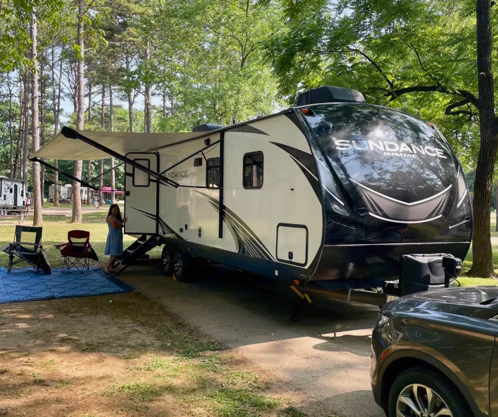 Sundance Ultra Lite RV rental setup at a shaded campground. Woman by steps, awning out, chairs & mat. Perfect for outdoor adventures!. Heartland RVs Sundance Ultra Lite 2021