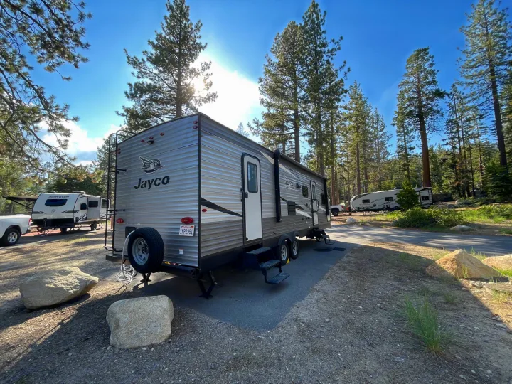 A Jayco travel trailer rv rental parked on asphalt at a sunny campground site, surrounded by tall pine trees.. Jayco Jay Flight SLX 2021