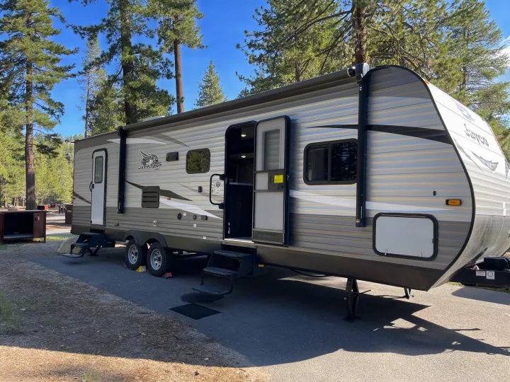 This grey Jay Flight rv rental travel trailer is set up at a sunny pine forest campsite with its door open.. Jayco Jay Flight SLX 2021