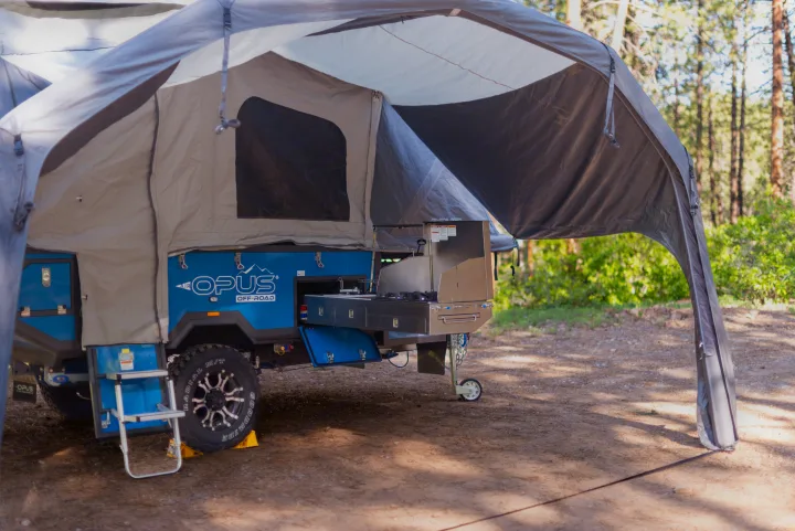 Blue OPUS Off-Road pop-up camper with outdoor kitchen setup at a scenic campsite. Book your RV rental adventure!. Opus OP4 2023