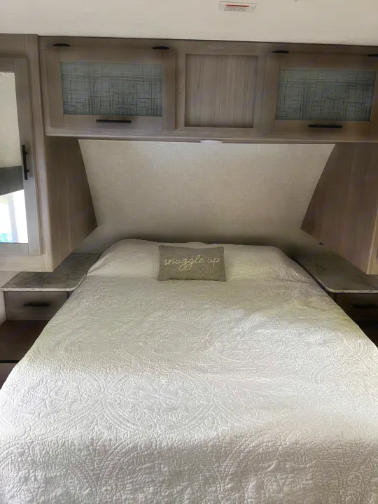Inside a cozy RV rental bedroom: a comfortable bed with a white quilt, overhead cabinets, and built-in nightstands.. Dutchmen Coleman Light 2021