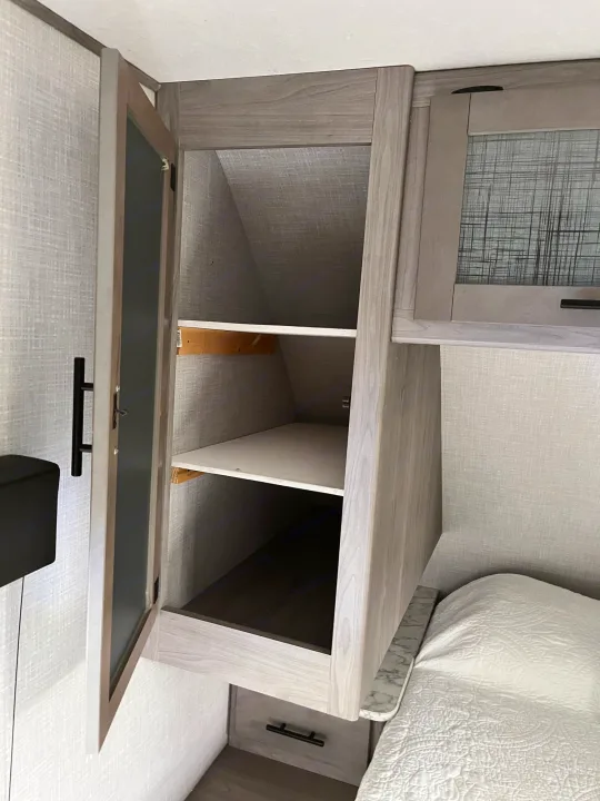 Inside an RV rental, discover smart bedroom storage: an open cabinet with shelves next to a cozy bed, optimizing compact living.. Dutchmen Coleman Light 2021