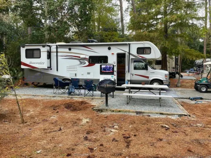 Spacious 'Sunseeker' rv rental parked in a wooded campground. Outdoor living area with chairs, grill, & picnic table.. Forest River Sunseeker 2016