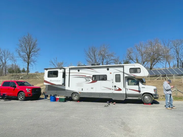 An RV rental, a white motorhome, and a red truck are parked on a sunny day. A man stands nearby, with solar panels in the background.. Forest River Sunseeker 2016