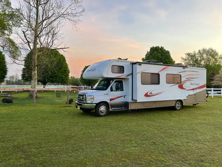 White Sunseeker RV rental parked in a grassy field with trees, a white fence, and a pig nearby.. Forest River Sunseeker 2016