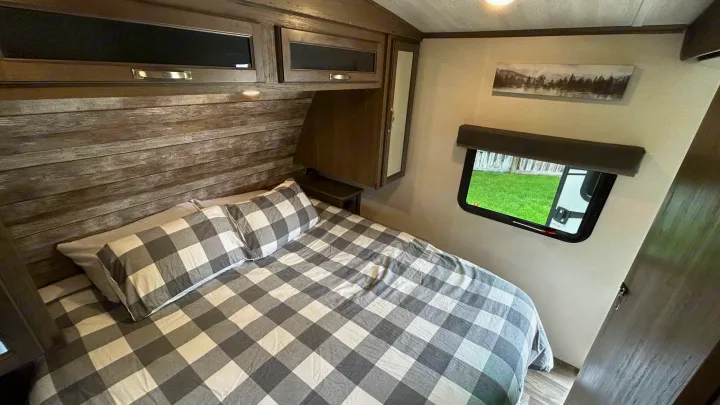 Inside an RV rental bedroom: a bed with gray buffalo check bedding, distressed wood wall, overhead cabinets, and side window.. Keystone Cougar Half-ton 2022