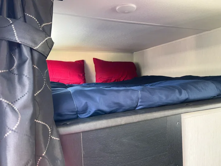 Cozy RV rental bunk bed: blue comforter, red pillows, light textured walls & dark patterned curtain. Ideal camper interior for rest.. Forest River Cherokee 2022
