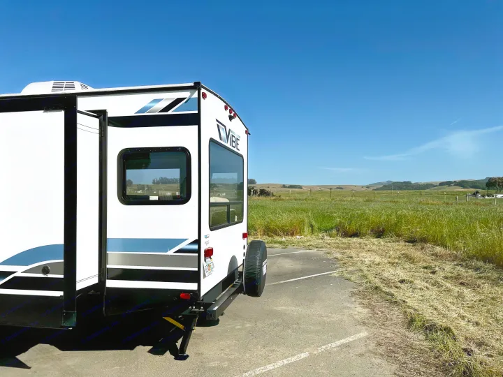A white VIBE **RV rental**, with an extended side, is parked beside a vibrant green field and hills under a clear blue sky.. Forest River Vibe 2022