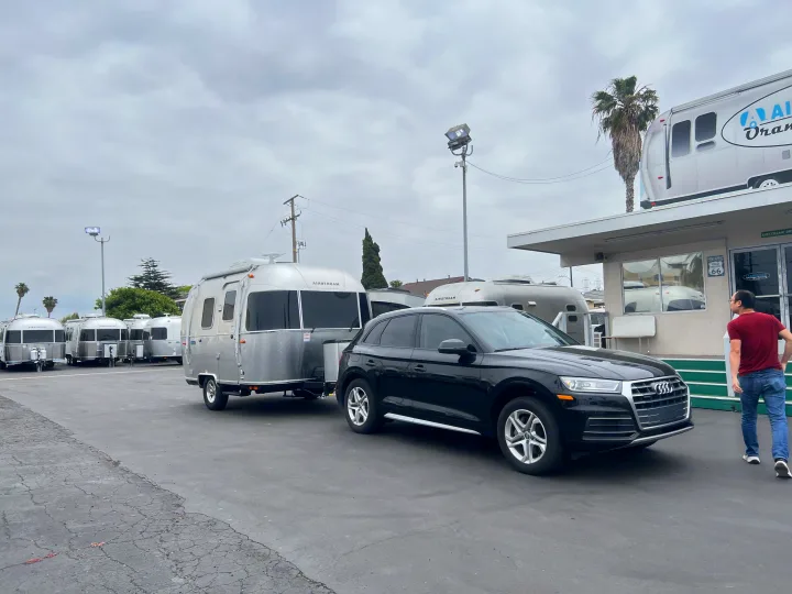 An Audi SUV is hitched to a silver Airstream travel trailer at an RV rental lot with more Airstreams visible.. Airstream Bambi 2023