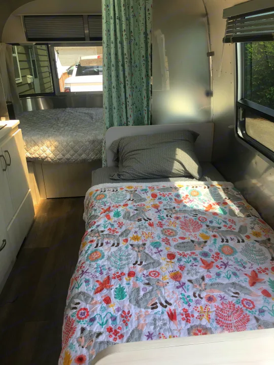 Bright rv rental interior, featuring a twin bed with a whimsical fox quilt and a larger bed beyond. Shiny walls reflect light.. Airstream Sport 2019