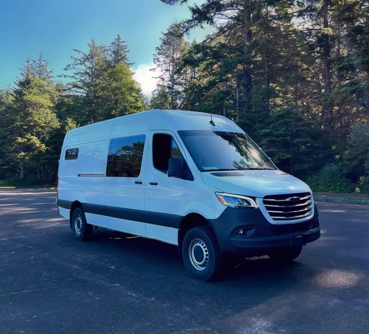 White Sprinter RV rental parked in a lot surrounded by lush green trees under a clear blue sky, ideal for travel adventures.. Freightliner Sprinter 2500 2019