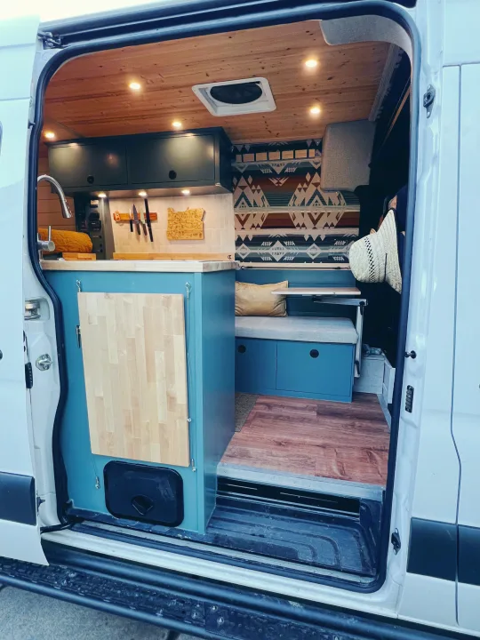Cozy customized camper van interior, perfect for rv rental. Shows kitchen, wooden ceiling, seating area, and patterned blanket.. Freightliner Sprinter 2500 2019