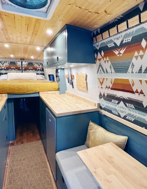 An RV rental's cozy interior, showcasing a bed, kitchen with blue cabinets, and a small dining area, all in a campervan.. Freightliner Sprinter 2500 2019