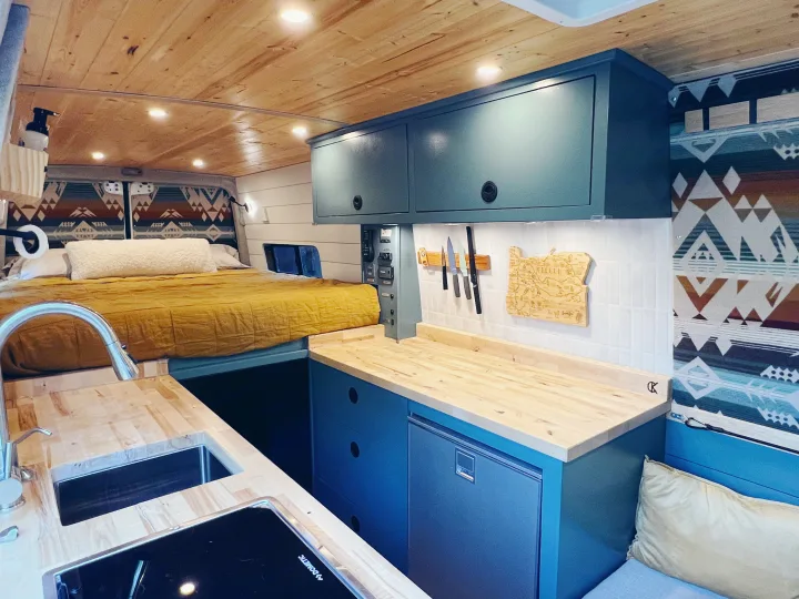 Inside a chic RV rental: a compact kitchen with blonde wood counters, teal cabinets, and a comfortable bed for van life.. Freightliner Sprinter 2500 2019