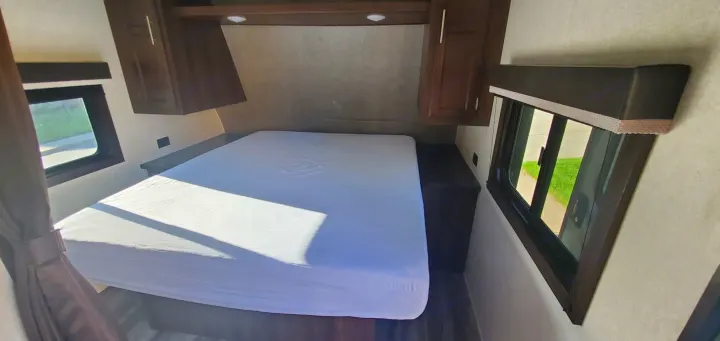 Bright RV rental bedroom featuring a full bed with white sheet, two windows, overhead cabinets, and built-in bedside storage.. Jayco Flight 2020