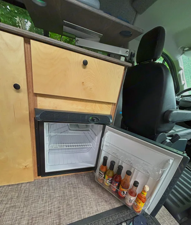 Compact RV rental kitchen with an open fridge showcasing various hot sauces, custom wood cabinetry, and a driver's seat.. Ford Custom Transit 2022