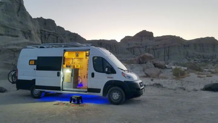 An RV rental camper van, brightly lit inside & with blue underglow, nestled against stunning desert mountains at dusk. Embrace van life travel!. Ram 3500 Extended 2021