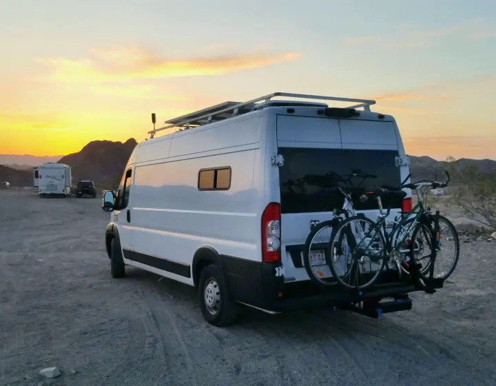 A white RV rental camper van with two bicycles on a rear rack, parked in a desert area with mountains at sunset.. Ram 3500 Extended 2021