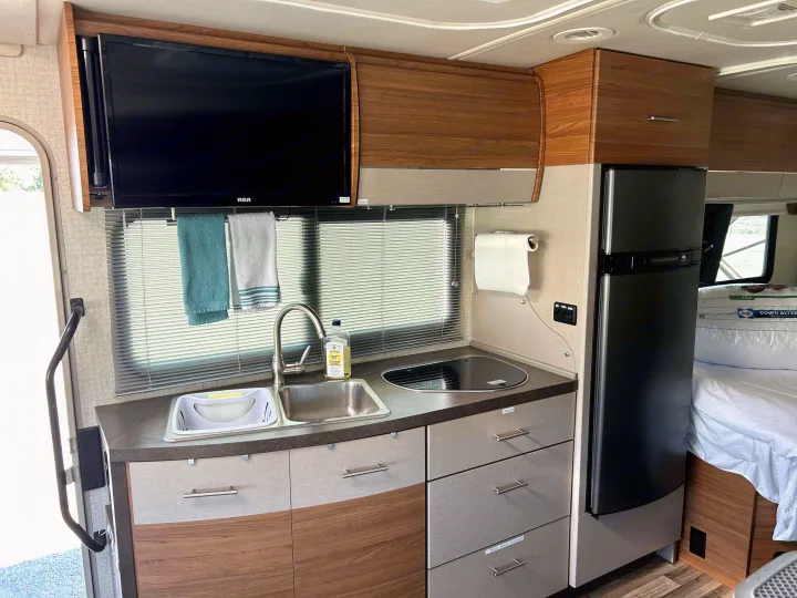 Functional RV rental interior with a compact kitchen featuring a sink, stovetop, fridge, overhead TV, and adjacent bed.. Winnebago View 2017