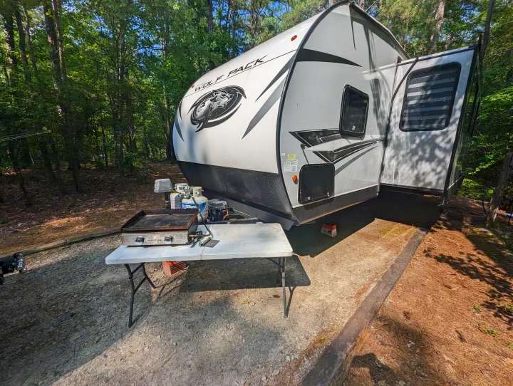 RV rental parked at a shady campsite. A "Wolf Pack" travel trailer with a griddle setup on a table for outdoor cooking.. Forest River Cherokee Wolf Pack Toy Hauler 2022