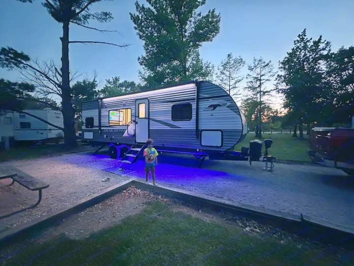 Enjoying our rv rental! A child stands next to a travel trailer illuminated by cool blue underglow lights at a peaceful campsite.. Heartland RVs Trail Runner 2022