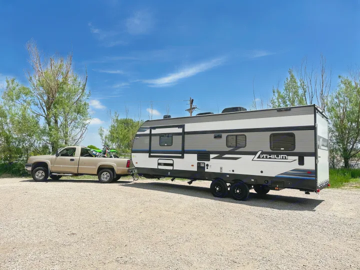 Tan pickup truck towing a sleek travel trailer RV rental, with a dirt bike in the bed, parked on gravel for outdoor adventures.. Heartland Lithium 2515S 2022