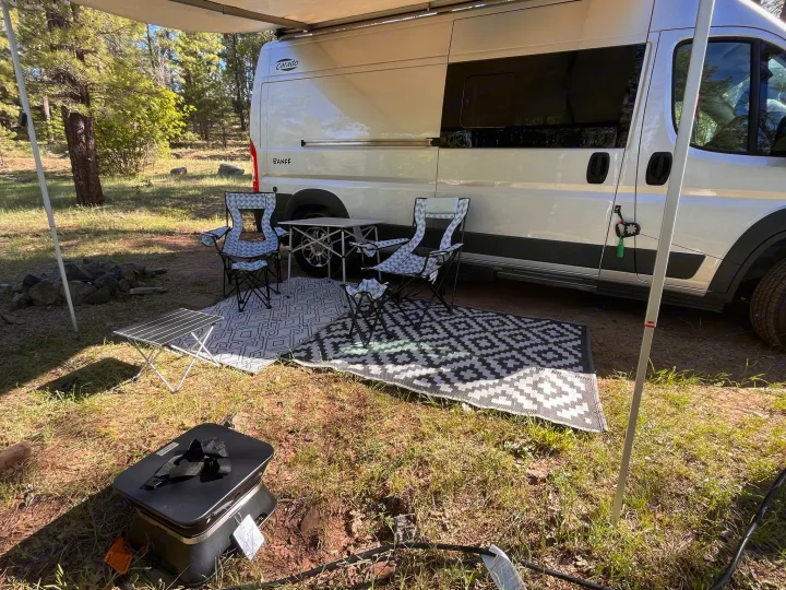 RV rental comfort: White camper van, extended awning, patterned rugs, camping chairs, table, and grill setup for outdoor living.. Carado Carado Class B 2018