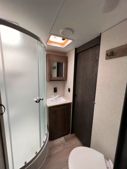 This RV rental bathroom features a curved, frosted glass shower, vanity with sink and mirror, and toilet, with wood-look flooring.. KZ Sportsmen LE 303KBLE 2021